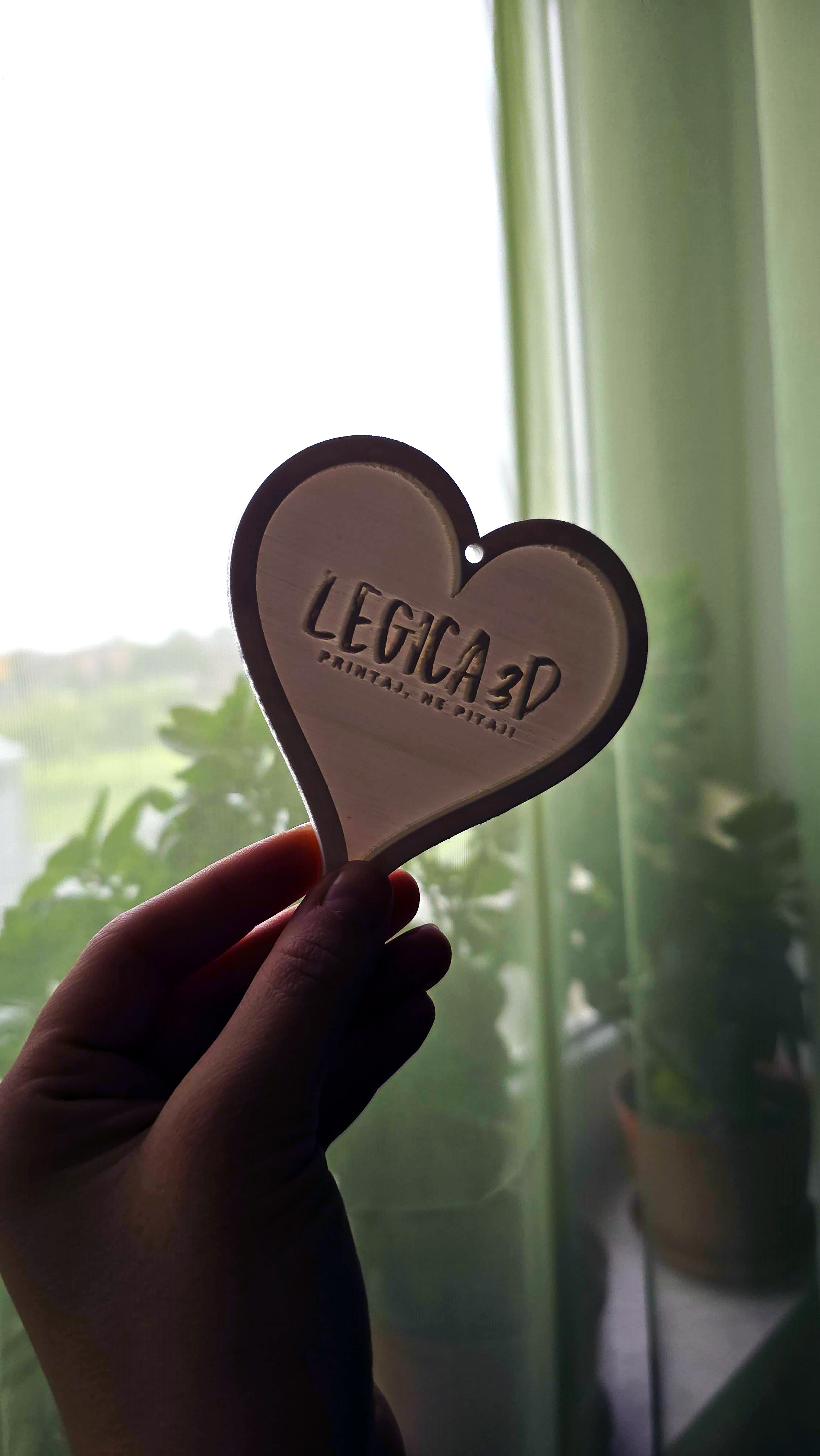 Ruka drži 3D printano srce s natpisom "Legica 3D – printaj, ne pitaj" ispred prozora kroz koji se vidi zelenilo i biljke.