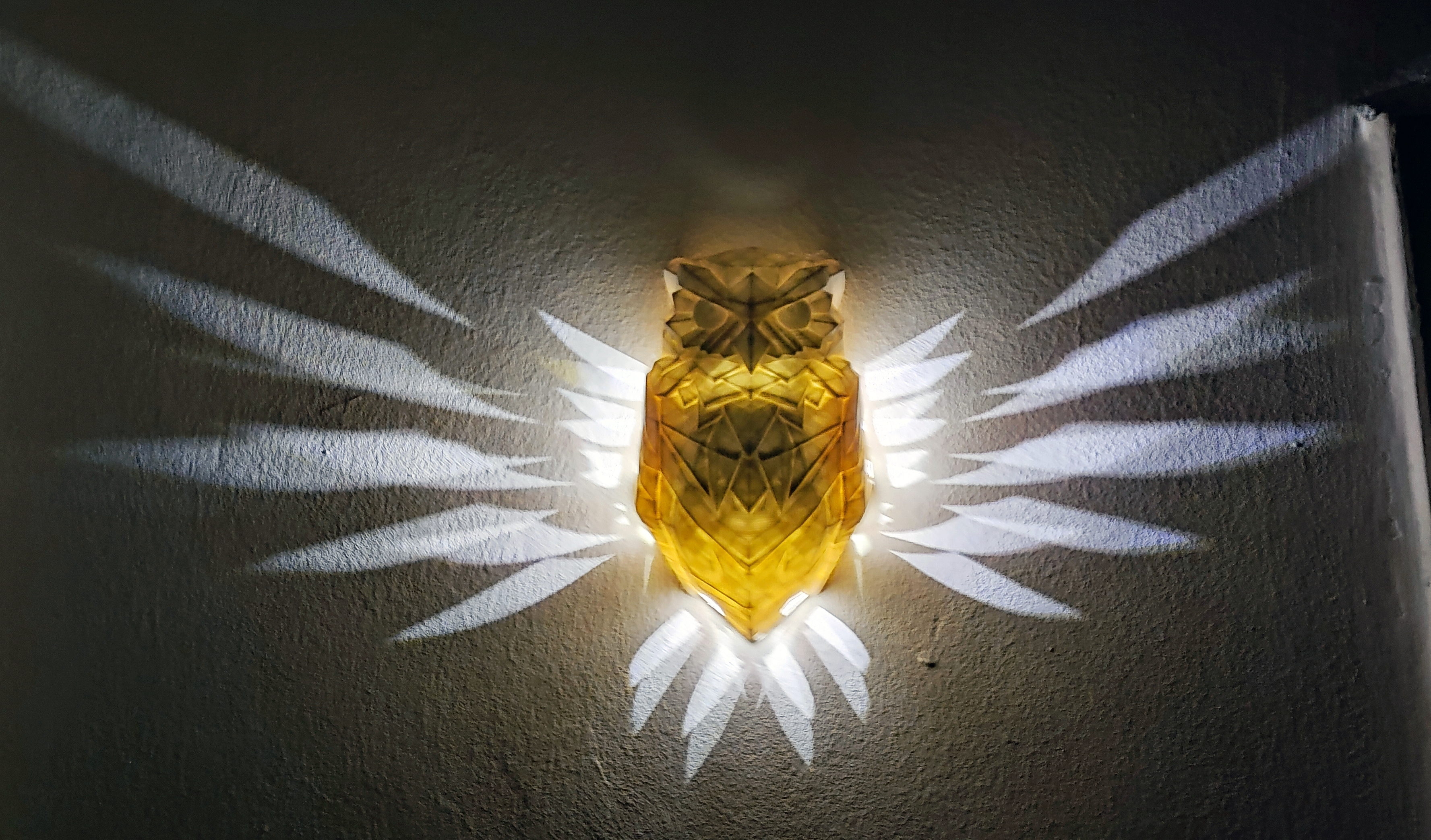 3D printana žuta lampa u obliku sovine glave, montirana na zid, s jedinstvenim svjetlosnim efektima koji stvaraju uzorak krila na zidu.